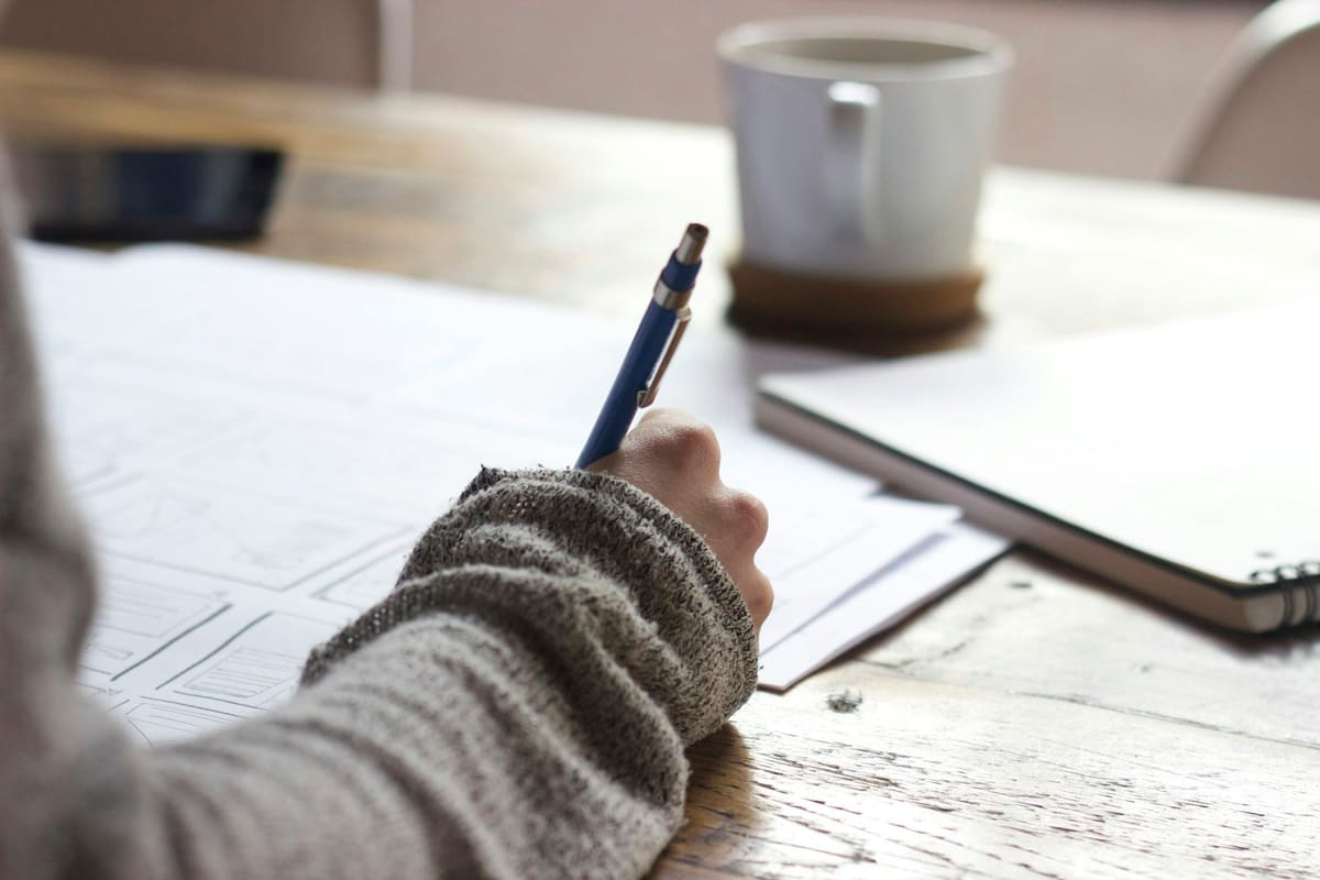 A person writing on printed sheets at a wooden desk, beside a coffee mug and a closed laptop