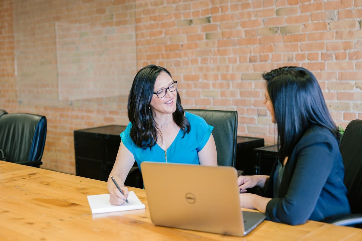 Two women talking across a wooden table, one taking notes beside an open laptop
