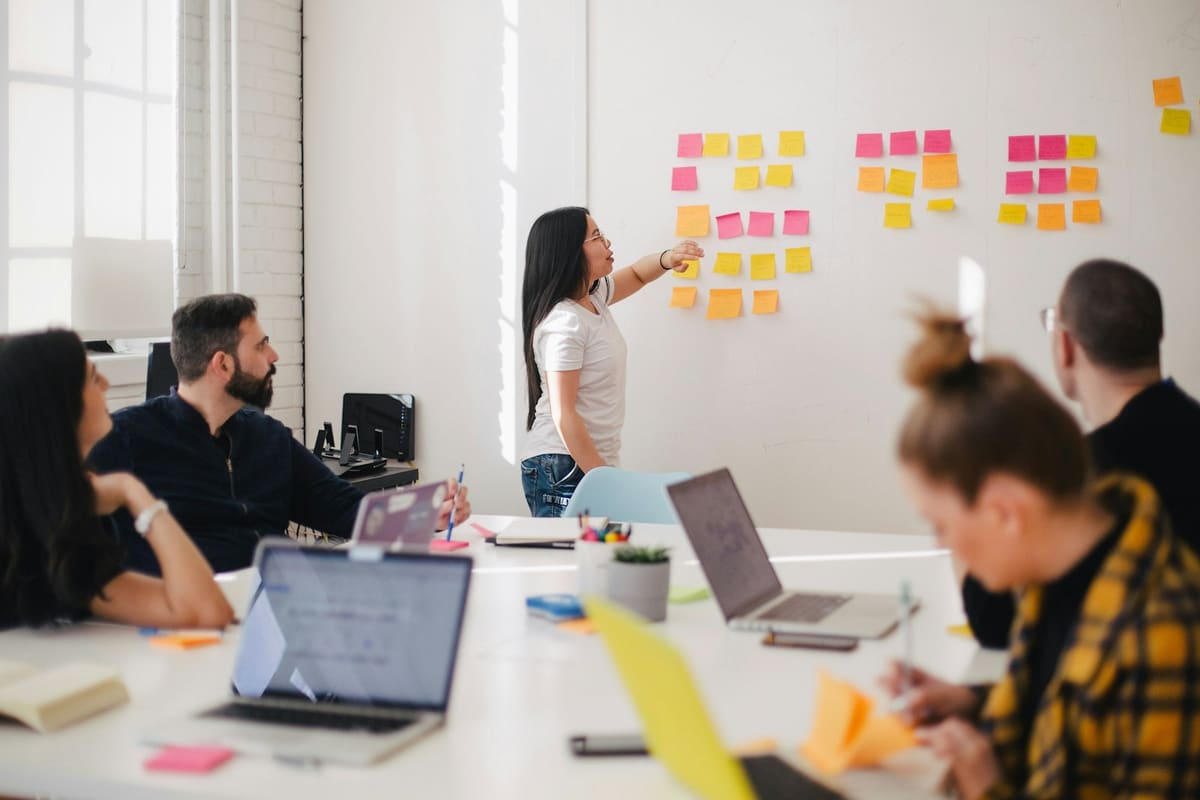 A woman pointing at rows of sticky notes on a white wall while colleagues with laptops watch from a table