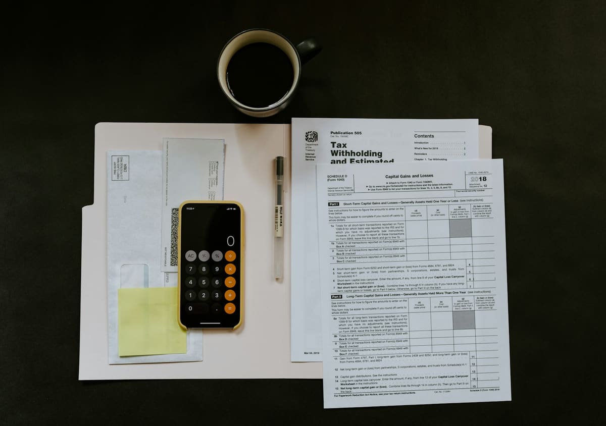 A phone calculator, a pen, a cup of black coffee and a folder of printed forms on a dark desk