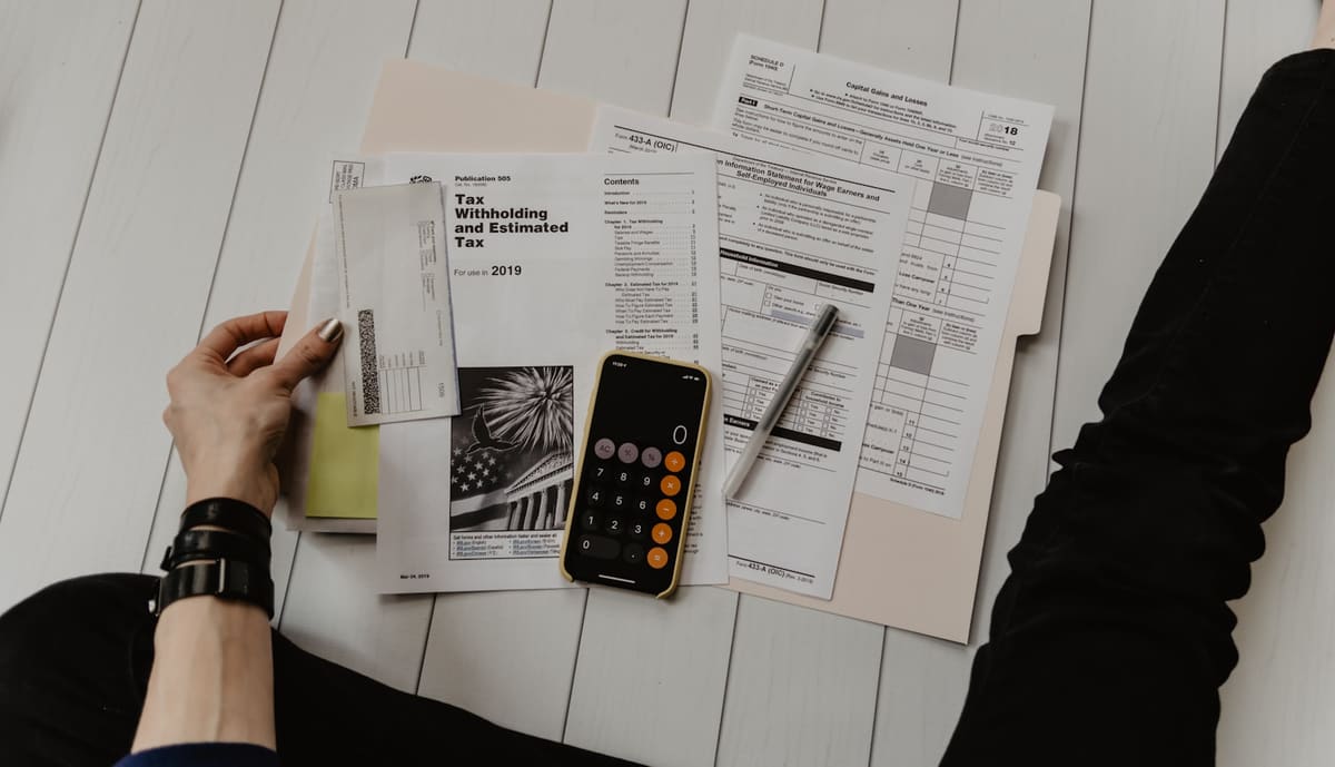 A person sorting printed forms and envelopes on a wooden floor, a phone calculator and a pen beside them