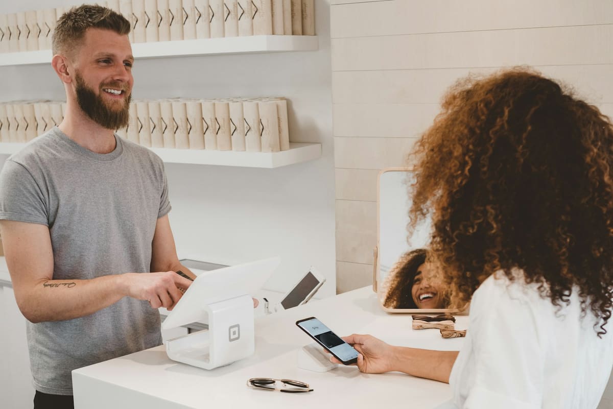 A smiling shopkeeper behind a white counter serving a customer who holds a phone