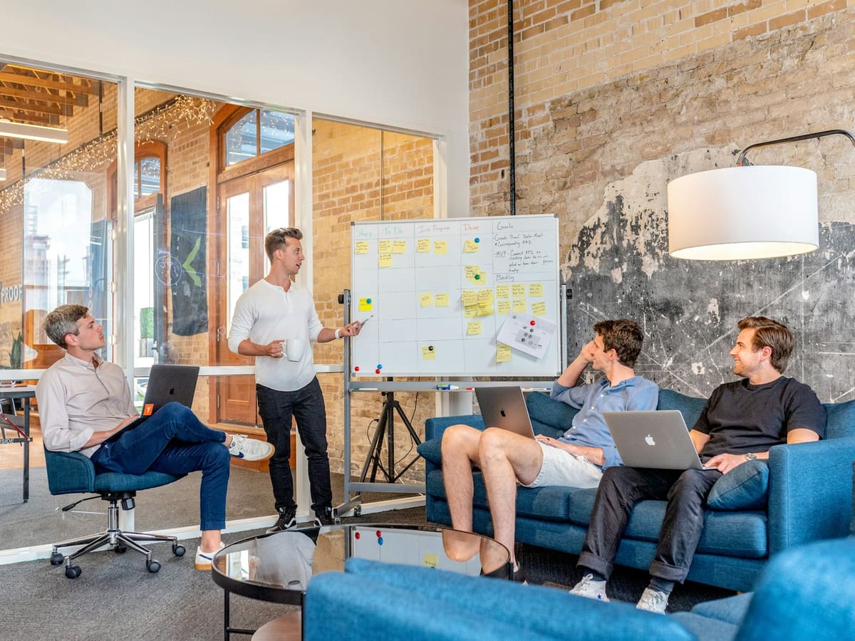 A man pointing at a whiteboard of sticky notes while three colleagues with laptops listen