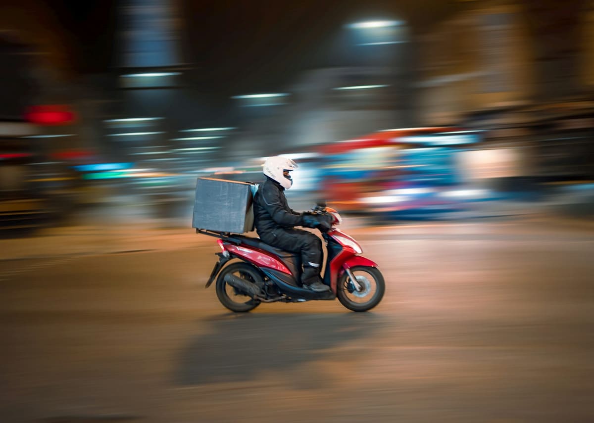 A rider in a white helmet on a red scooter with a box on the back, blurred city lights behind