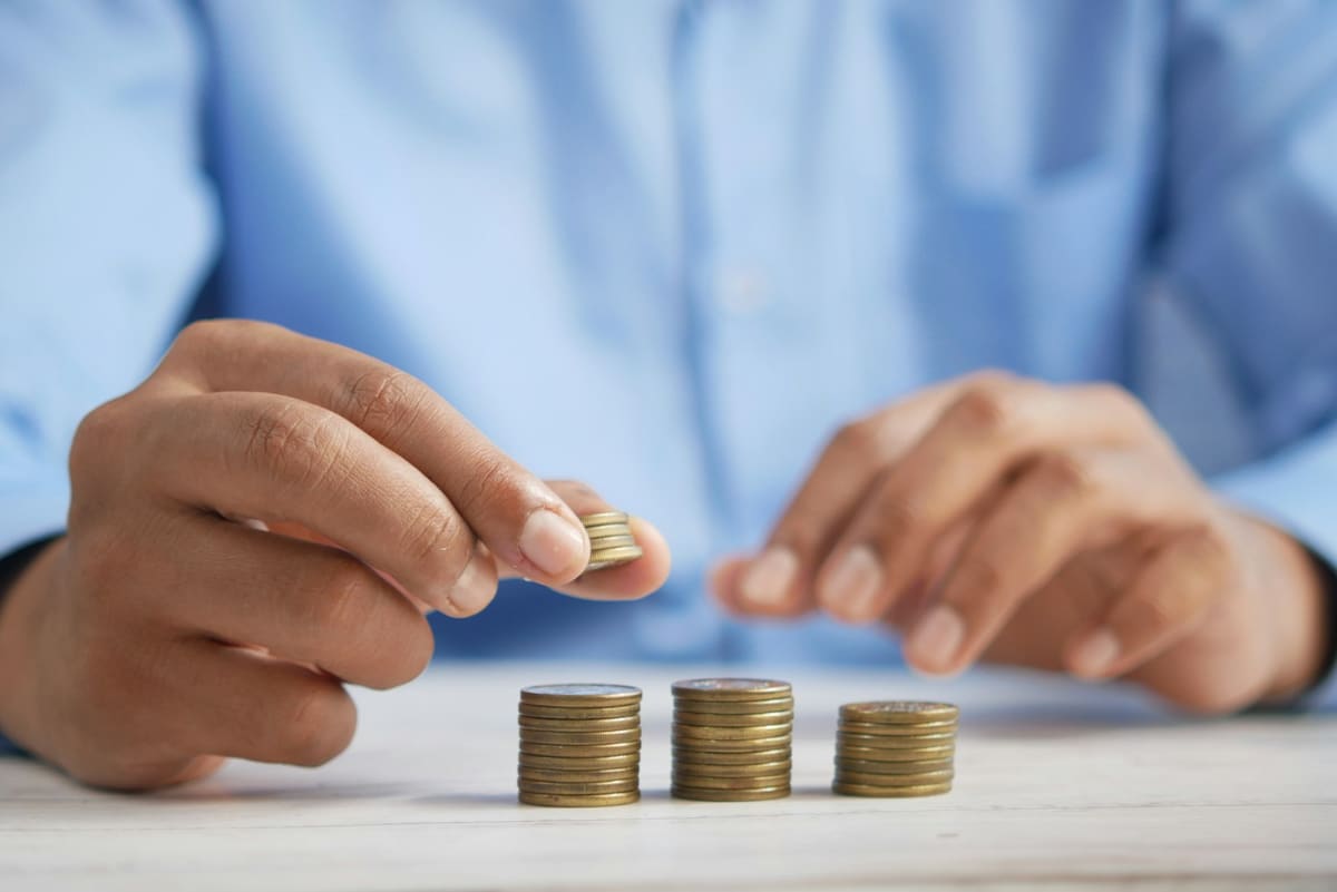 A hand holding a coin beside three short stacks of coins on a table