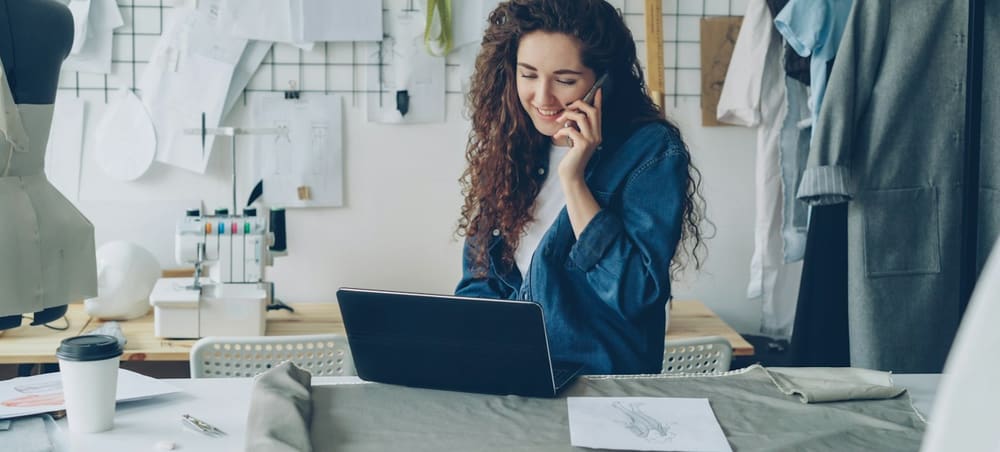 A woman in a tailoring workroom, on the phone with a laptop open in front of her.